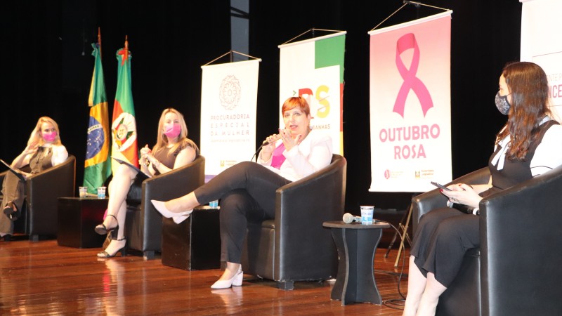Ana Costa fala enquanto outras três mulheres ouvem. Todas estão sentadas em poltronas em um palco com banners e bandeiras do RS e do Brasil. 