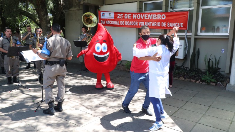 Um homem dança com uma profissional de saúde. Um boneco com fantasia em formato de gota de sangue também dança ao som da Brigada Militar, que também aparece na imagem. 