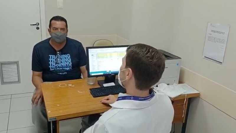 Paulo Roberto Ferreira Costa e Serlio Zanol conversam sentados, separados por uma mesa, num consultório médico. Os dois estão de máscara.