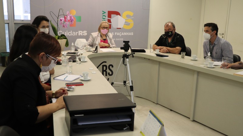 Secretárias Arita e Ana Costa participam de uma reunião com mais quatro pessoas (duas mulheres e dois homens). Todos estão de máscara e sentados atrás de uma mesa em formato de U. 