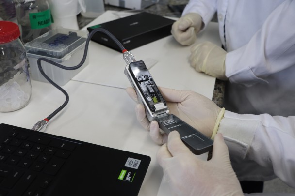 A imagem mostra as mãos de uma profissional de saúde manuseando o equipamento que realiza o processo de sequenciamento genômico completo. 