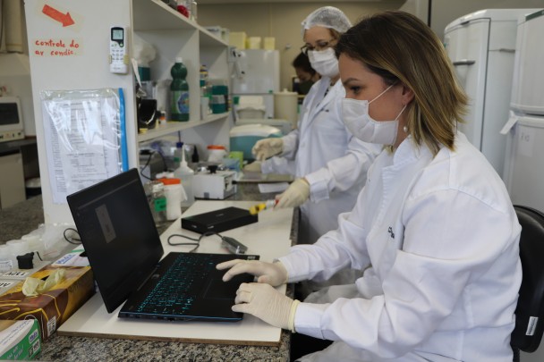 Uma profissional da saúde trabalha com um computador. Outras duas manuseiam frascos e equipamentos. 