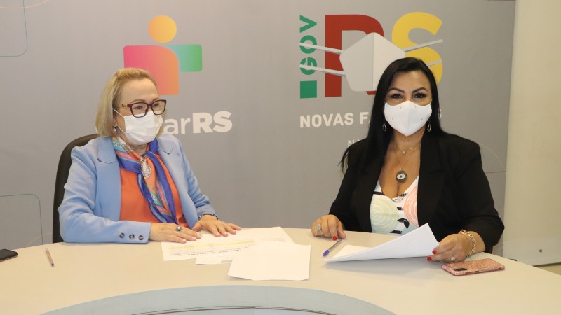 Secretária Arita e Kelly, sentadas atrás uma mesa e de máscara, sorriem. Kelly segura um documento.  