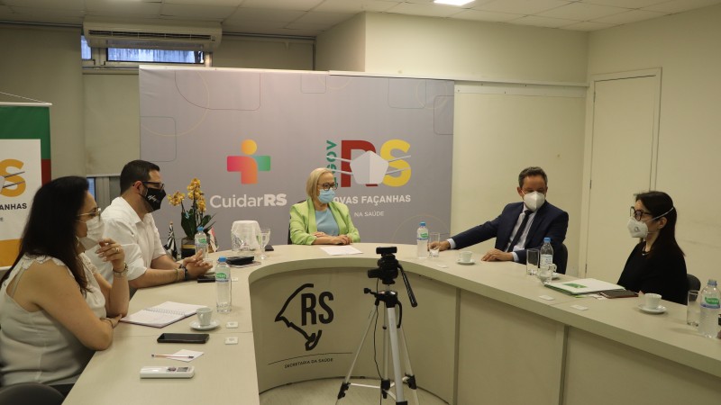 A imagem mostra a reunião com a Secretária Arita; o prefeito e mais três pessoas. Os cinco estão de máscara e sentados atrás de uma mesa em formato de U. 