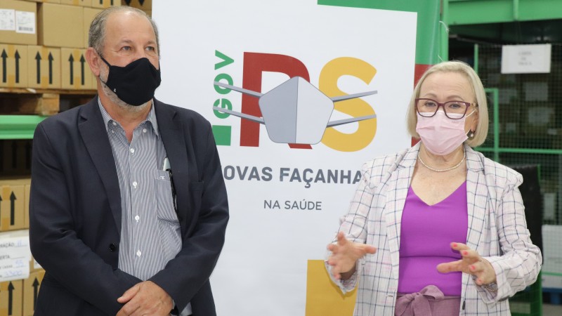 Ao lado do secretário José Luiz Stédile, Arita Bergmann fala e gesticula no Almoxarifado. Os dois estão de pé e de máscara. Atrás dele, há um banner com o logotipo da Secretaria Estadual da Saúde. 
