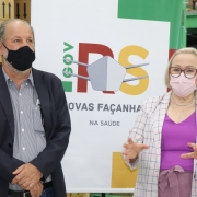 Ao lado do secretário José Luiz Stédile, Arita Bergmann fala e gesticula no Almoxarifado. Os dois estão de pé e de máscara. Atrás dele, há um banner com o logotipo da Secretaria Estadual da Saúde. 