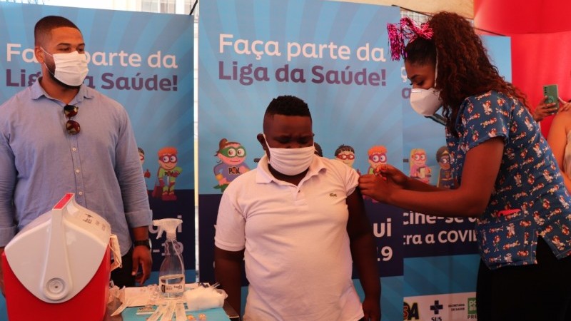 Uma mulher aplica vacina em Dionatan. Do lado dos dois, um adulto. Todos estão de máscara. Atrás deles, tem banners da campanha.