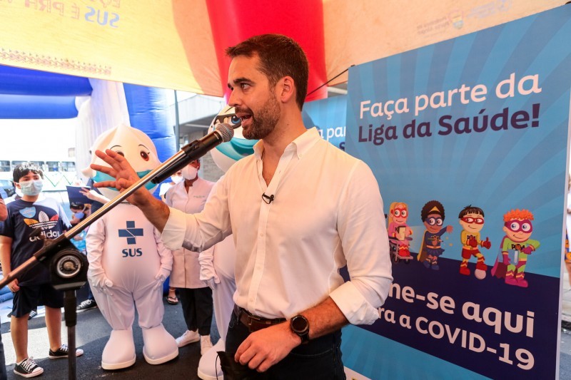 Governador Leite discursa tendo, do lado, o Zé Gotinha, uma profissional de saúde e uma criança. Atrás deles, banners da campanha.