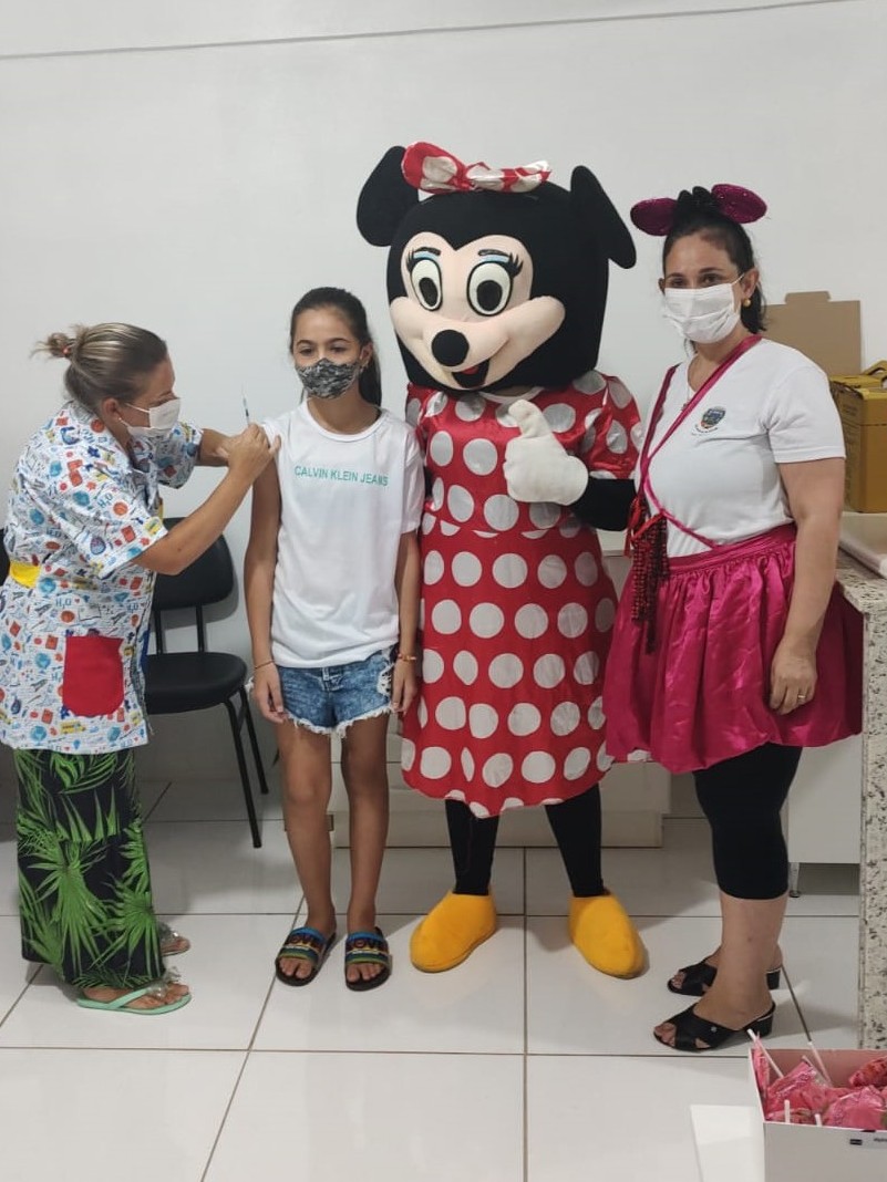 Uma profissional de saúde fantasiada vacina uma menina que está do lado da Minnie e de outra mulher fantasiada. 