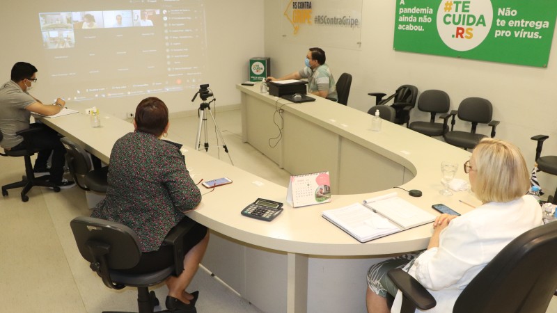 As Secretárias Arita e Ana Costa e mais dois homens veem a live que aparece no telão. Os quatro estão sentados atrás de uma mesa em formato de U.  