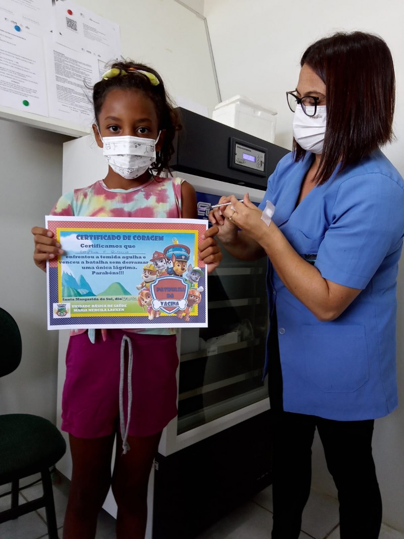 Uma menina segura o seu certificado de coragem enquanto é vacinada por uma profissional de saúde. As duas estão de pé e de máscara. Atrás delas, tem uma câmara fria.  