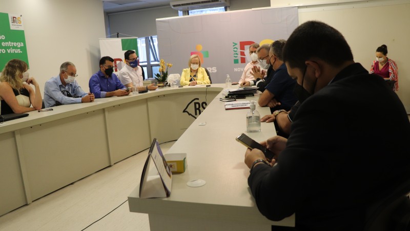 Os vários participantes de reunião, entre eles a Secretária Arita, estão sentados atrás de uma mesa em formato de U. 