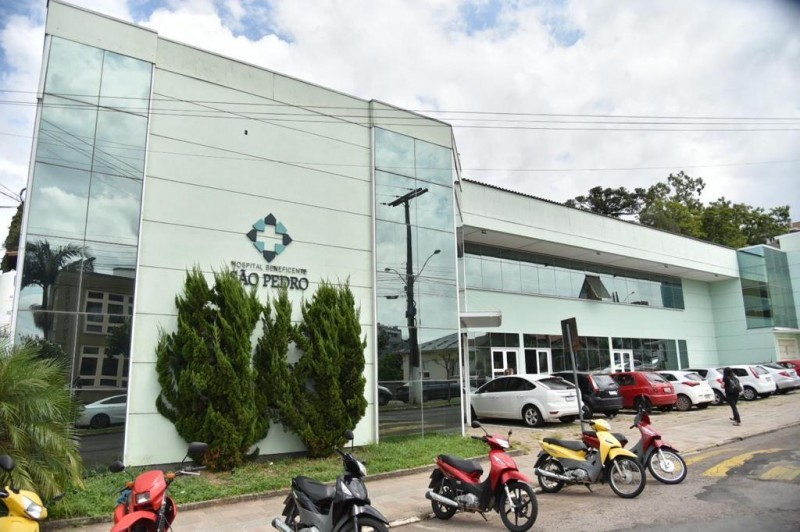 Imagem da parte externa do hospital com carros e motos estacionados.