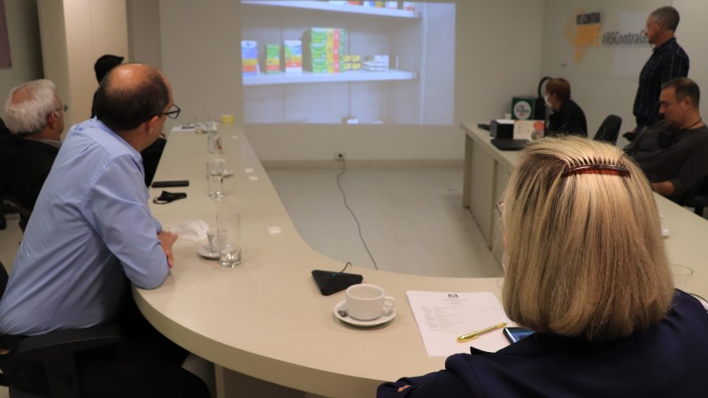 Os participantes da reuni&atilde;o, entre eles a Secret&aacute;ria Arita, assistem o v&iacute;deo que aparece no tel&atilde;o. Todos est&atilde;o sentados atr&aacute;s de uma mesa em formato de U. 