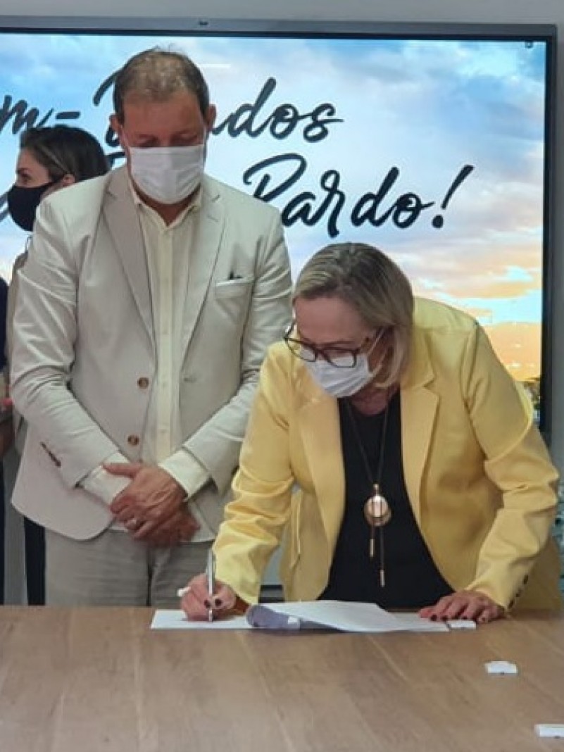 Abaixada, a Secretária Arita assina o documento. O prefeito Edivilson observa. Os dois estão de máscara.