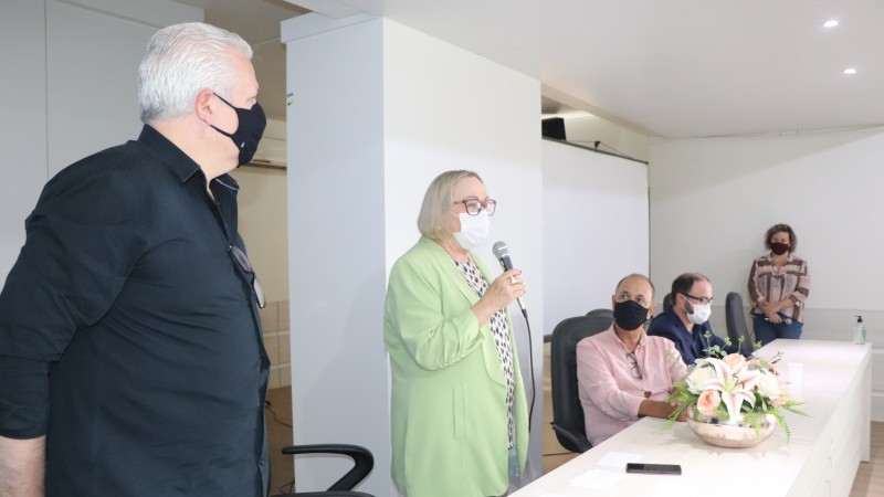 De pé, a Secretária Arita segura o microfone e discursa. Ao seu lado, à esquerda, atrás da bancada, um homem, também de pé. À direita, outros dois homens, sentados. No fundo, a mestre de cerimônias está de pé. Todos estão de máscaras.   
