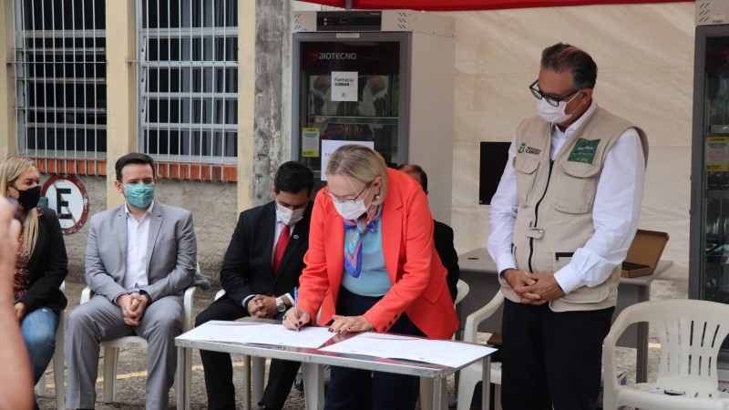 Abaixada, a Secretária Arita assina um documento. Ao seu lado, o prefeito de Canoas. Ao fundo, quatro pessoas sentadas. Todos estão de máscara. A foto foi tirada na parte externa da  Divisão de Suprimentos da Secretaria da Saúde, em Porto Alegre. 