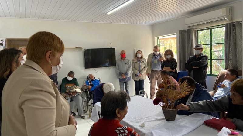 De pé, Ana Costa observa os moradores e outras pessoas que estão no residencial, alguns estão sentados, outros estão de pé.