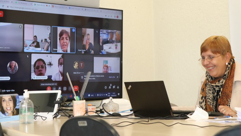 Ana Costa na reunião centros TEAcolhe sorrindo em frente ao computado e ao lado de um monitor com demais participantes online