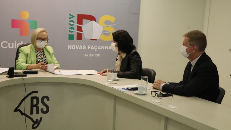A Secretária Arita manuseia alguns documentos enquanto uma mulher e um homem observam. Os três estão de máscara e sentados atrás de uma mesa em formato de U.  