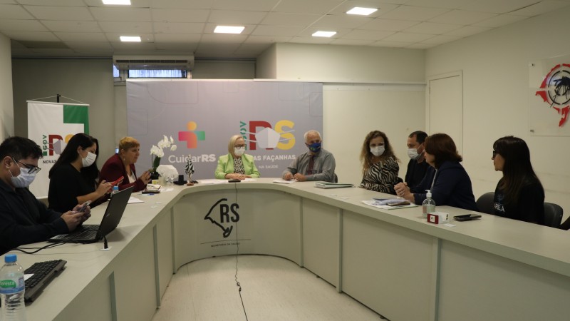 Nove pessoas em reunião na sala de monitoramento, incluindo as secretárias Arita Bergmann e Ana Costa. Todos estão sentados atrás de uma mesa em formato de U. 