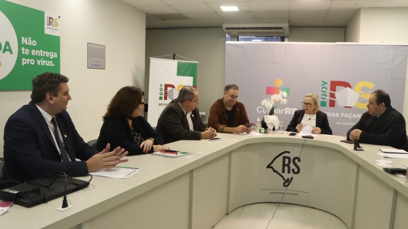 A Secretária conversa com os participantes da reunião (uma mulher e quatro homens). Todos estão sentados atrás de uma mesa em formato de U. 