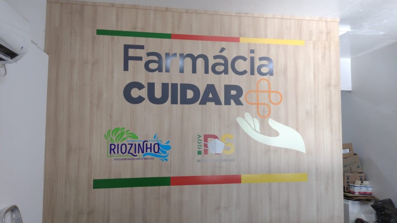 A imagem mostra uma parede com os logotipos do Farmácia Cuidar + e do Governo do Estado.