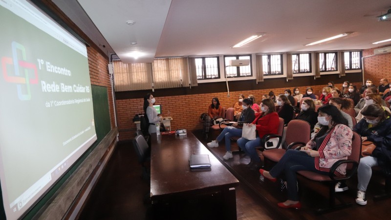 Auditório com público, uma pessoa ao lado de uma mesa de madeira falando aos presentes