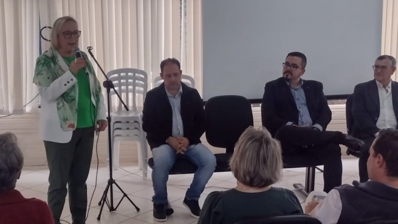 De pé e segurando o microfone, a Secretária Arita discursa. À direita, três homens sentados. À frente, a plateia está sentada.