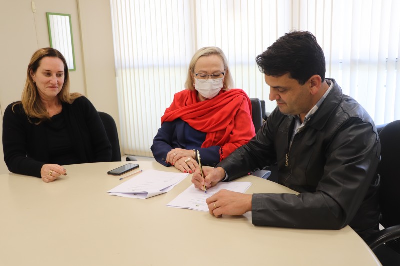 À direita, Marcelo assina um documento. A Secretária Arita e Ana Baggio observam. Os três estão sentados atrás de uma mesa circular. 
