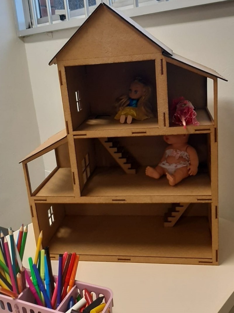 A imagem mostra uma casinha de madeira com 3 andares. Duas bonecas estão dentro da casa. Na frente da casa, uma caixa com vários lápis de cor. 