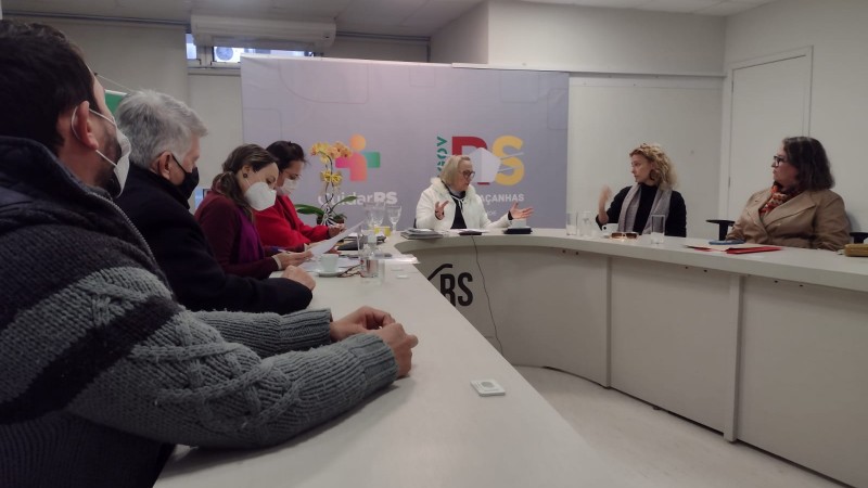 A Secretária Arita fala enquanto os seis participantes da reunião (quatro mulheres e dois homens). Todos estão sentados atrás de uma mesa em forma de U.