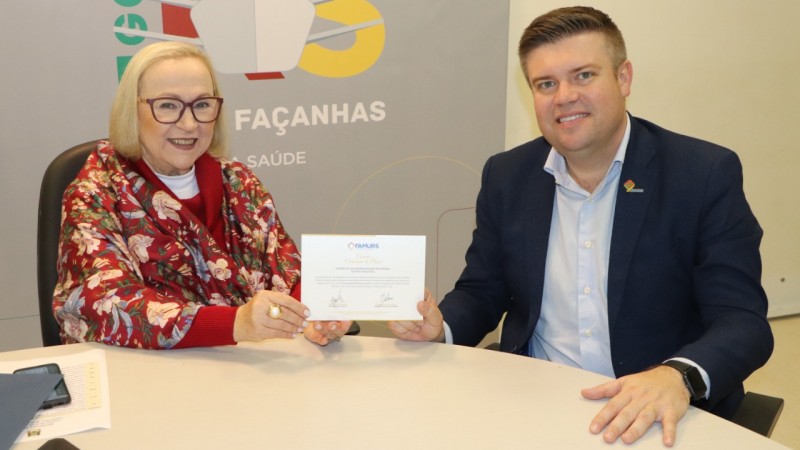 Sentados atrás de uma mesa circular, a Secretária Arita e o Prefeito seguram, juntos, o convite.