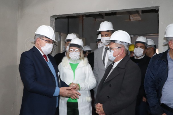 A Secretária Arita e alguns outros homens estão de pé, de capacete branco na parte interna das obras.   