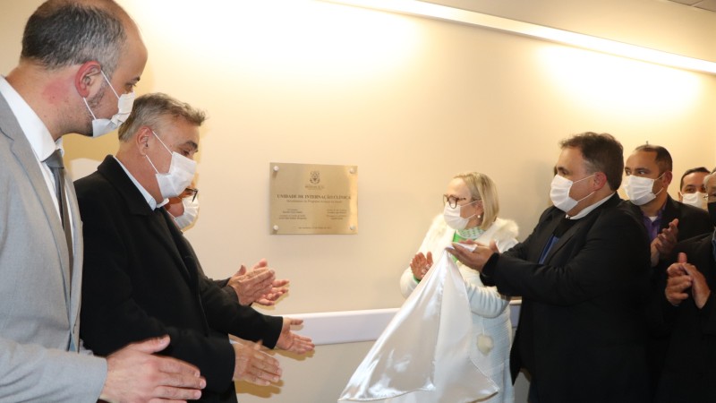 A Secret&aacute;ria Arita e outro homem seguram o pano branco. Outros homens aplaudem o descerramento da placa do hospital. 