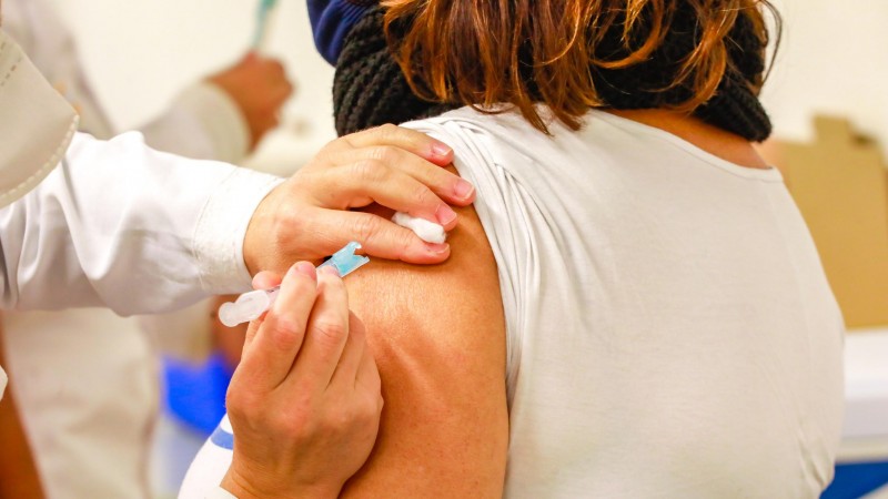 A imagem mostra as duas mãos de uma profissional de saúde vacinando o braço de uma mulher. 