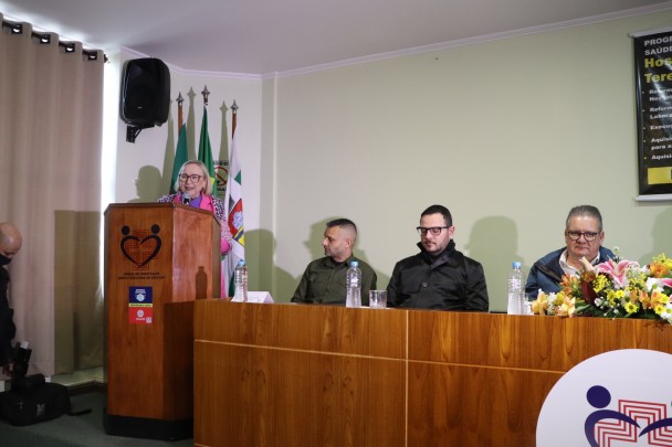 De pé e atrás de um púlpito, a Secretária Arita discursa. Do lado direito, sentados atrás da bancada, o Governador Ranolfo e dois homens. 