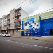 A imagem mostra a parte externa do prédio da Farmácia. Do lado esquerdo, tem outro prédio e alguns carros estacionados. 