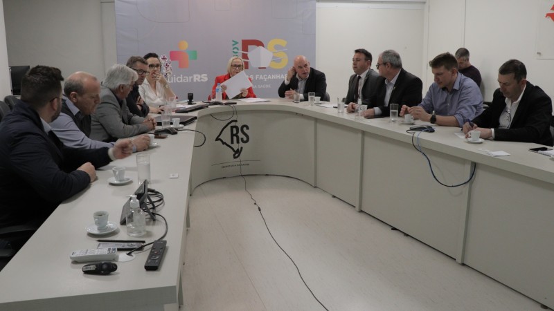 A Secretária Arita e mais dez pessoas (uma mulher e nove homens) estão sentados atrás de uma mesa em fomati de U e conversam.  