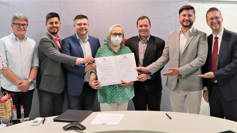 Secretária Arita Bergmann, em pé, mostrando os documentos assinados, junto com os participantes da cerimônia de assinatura do convênio com o Hospital de Restinga Seca