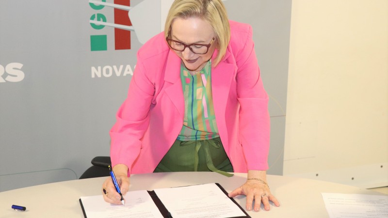 Abaixada, a Secretária Arita sorri e assina um documento.