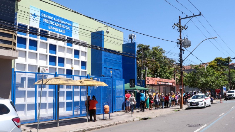 A imagem mostra a parte externa do Centro Logístico de Medicamentos Especiais com uma fila de pessoas esperando na rua e três carros. 