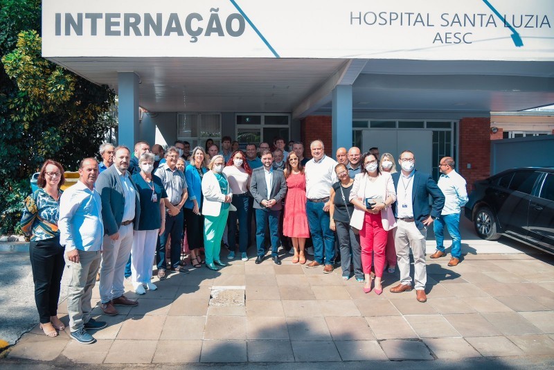 Um grupo com dezenas de pessoas em pé no saguão em frente ao Hospital que tem um placa com o nome Hospital Santa Luzia