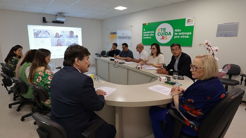 Secretária Arita Bergmann está sentada com mais 10 pessoas (4 mulheres e 6 homens) ao redor da mesa em formato oval do gabinete. Um painel com uma vídeoconferência está online na parede.

 