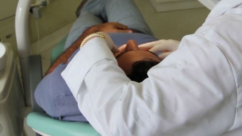 Atendimento em Saúde Bucal aparece o braço de uma dentista com as mãos com luvas examinando um paciente 

