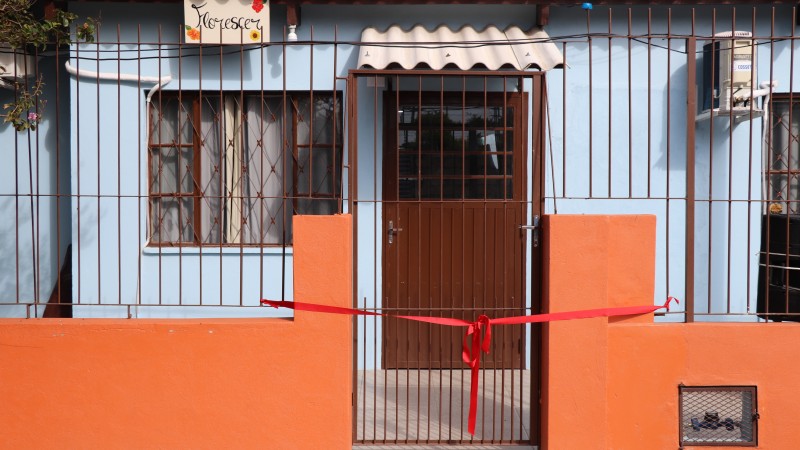 Foto da frente do residencial, com fita vermelha na frente do portão. O muro da casa é laranja, a casa é azul claro, as grades e portas são na cor marrom. No lado superior esquerdo está uma placa escrito "Florescer".