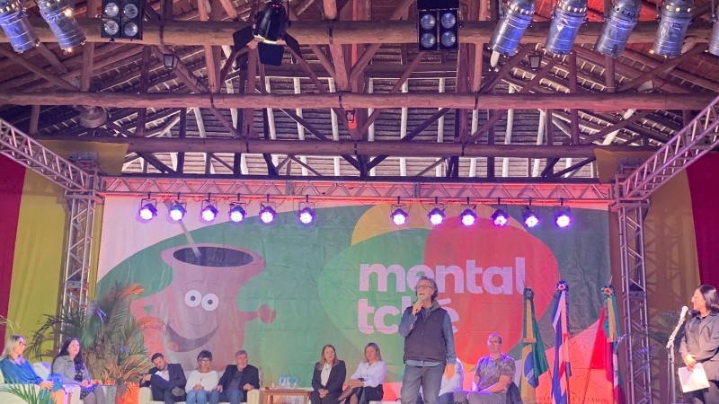 Um palco com faixa do evento e várias pessoas sentadas e um homem falando ao microfone