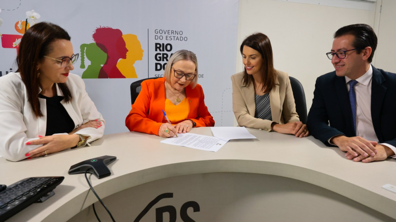 Secretária Arita assina portaria na mesa de reuniões do gabinete, acompanhada da prefeita e demais autoridades. Arita veste uma blusa e um blazer laranja. Ao fundo está o logo do governo do estado.