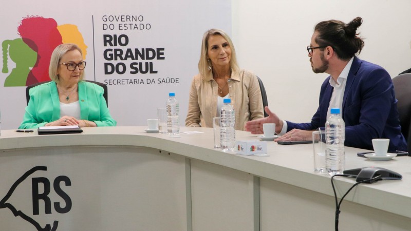 Na sala de reuniões do Gabinete, Arita Bergmann está sentada vestindo um blazer verde. Ao lado, a presidente da FAURGS, Ana Rita Facchini, vestindo camisa bege. Ambas prestam atenção no pesquisador Eduardo Zimmer, que está falando sobre a importância do convênio assinado. Zimmer veste um terno azul marinho, usa óculos e tem os cabelos castanhos compridos, presos em um coque. As três pessoas citadas na foto são brancas. Ao fundo, o banner com o logo do do governo do estado do rio Grande do Sul.