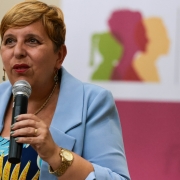 Secretária Ana Costa fala ao microfone. Ela veste um blazer azul claro, tem cabelos loiros e curtos. Ao fundo, desfocado, está o banner do Governo do Estado.